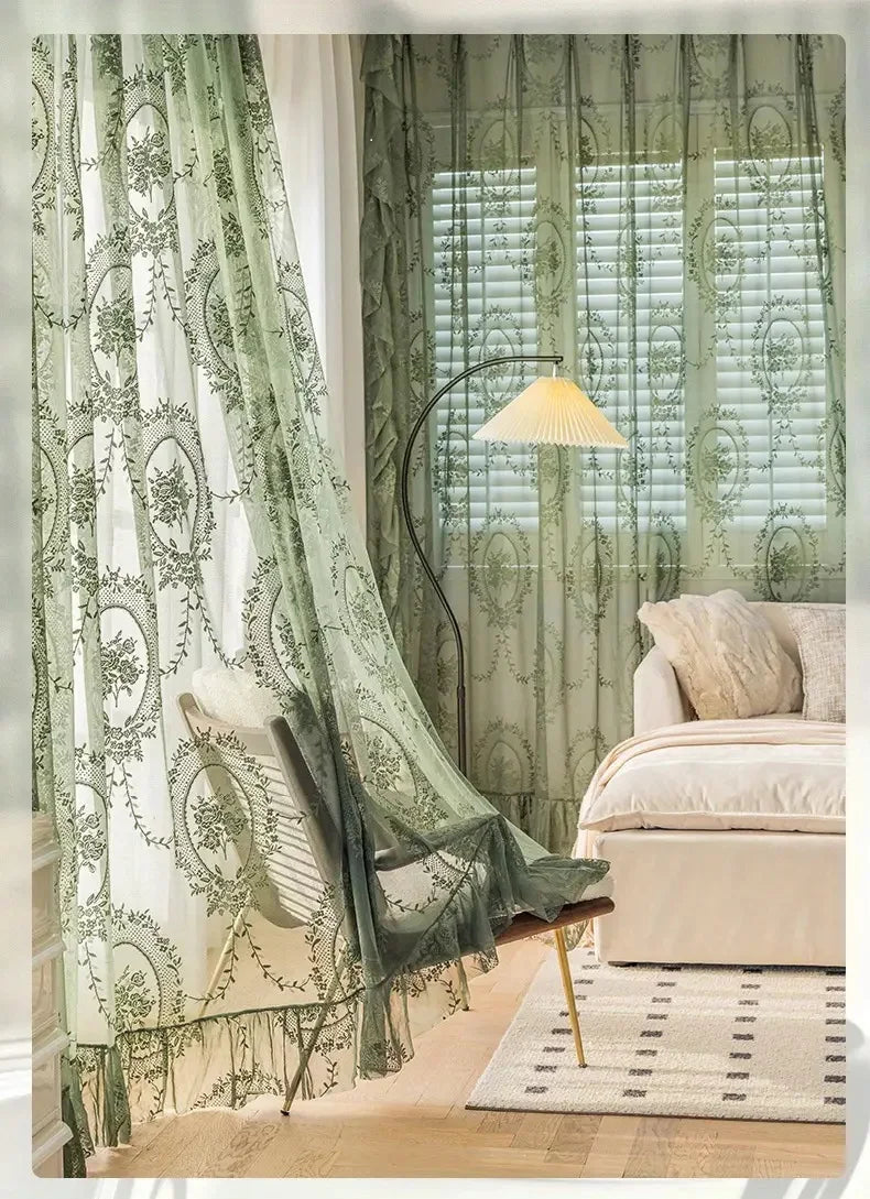 A sheer floral lace curtain panel in sage green hangs next to a window with slatted blinds and a modern lamp.