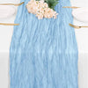 A light blue cheesecloth table runner with ruffled texture, set for a meal with white plates and gold cutlery.