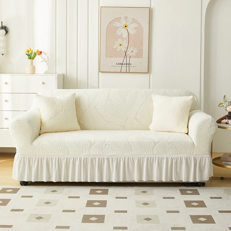 A cream-colored Jacquard elastic sofa cover with a pleated skirt displayed in a bright, modern living room.