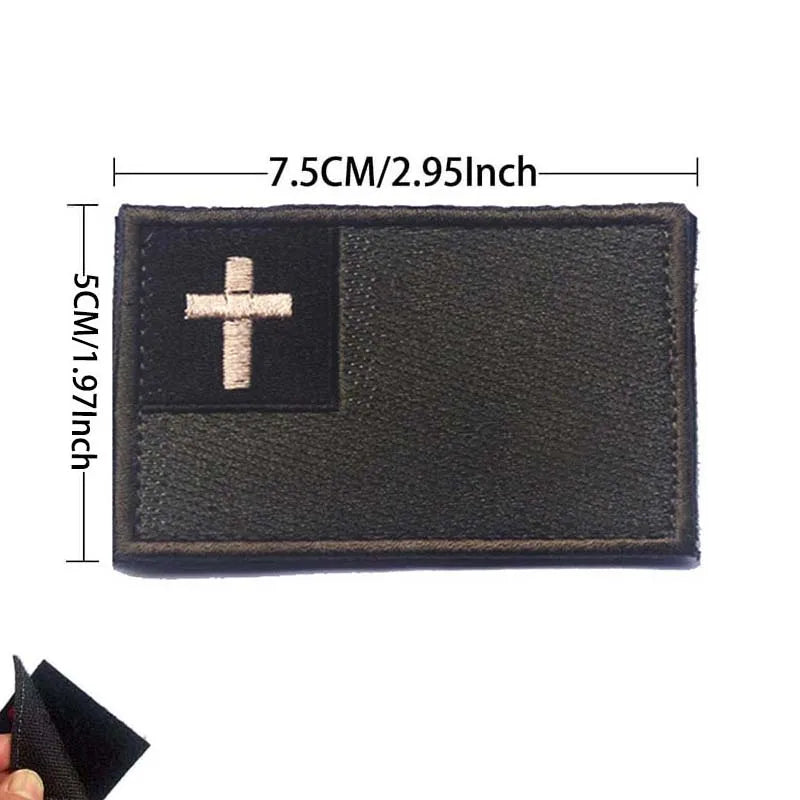 A rectangular dark brown embroidered patch featuring a white cross on a black background.