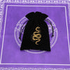 A black velvet tarot cards storage pouch with a gold coiled snake design rests on a purple patterned surface.