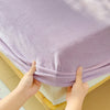 A hand is holding up a purple flannel fitted sheet with deep pockets, showing the fabric texture and elastic edge.