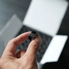 A hand holds a black embroidery bobbin thread against a blurred background of a laptop.