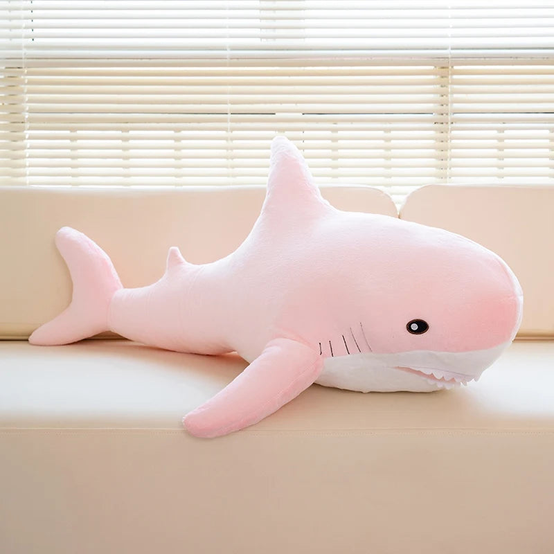 A pink giant shark plush toy lying on a light-colored surface near a window.