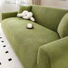 A moss green stretch jacquard sofa slipcover in a textured ribbed fabric with a white decorative pillow and a cup on the seat