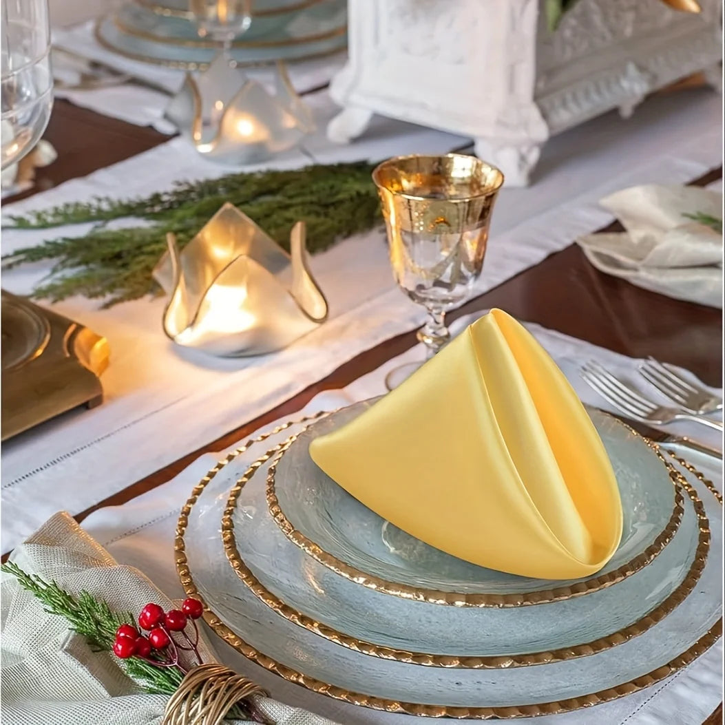 A set of silky yellow satin dinner napkins is displayed on a table with gold accents and greenery.