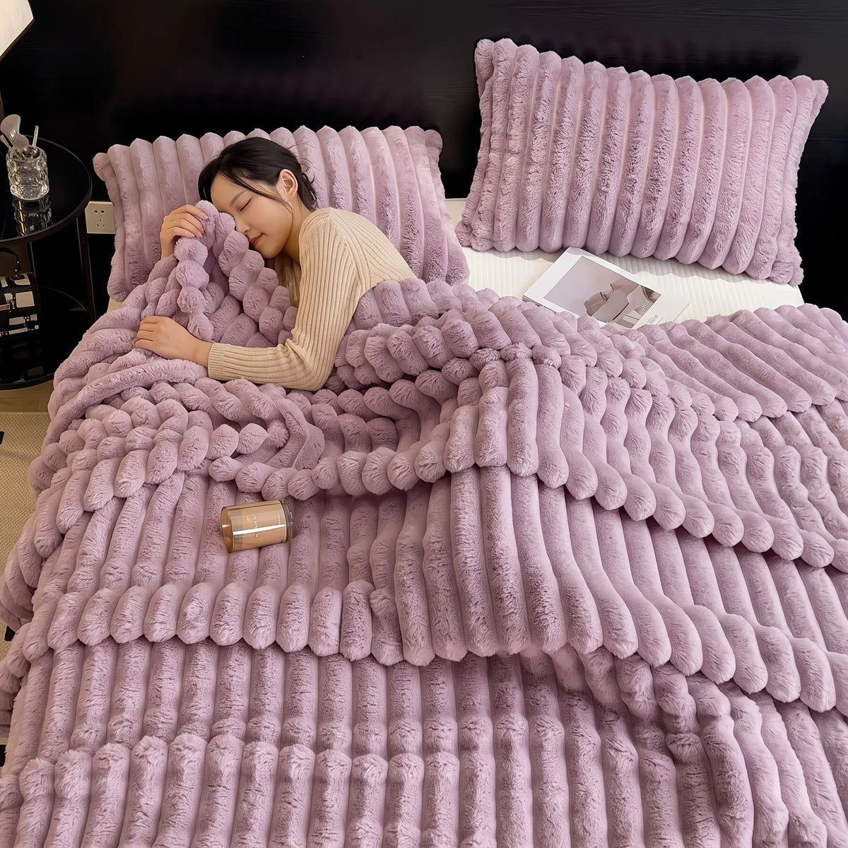 A woman is relaxing under a thick, fluffy mauve fleece throw blanket in a bedroom setting.