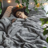 A woman is sleeping under a soft gray flannel throw blanket with green foliage framing the shot.
