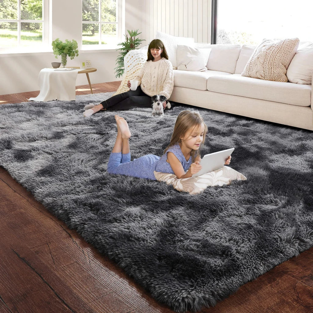 A young girl lies on a dark gray fluffy floor mat in a bright living room with a woman and a dog nearby.