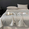 A light gray lightweight cooling blanket is displayed on a bed with a dark gray wall and white pillows.