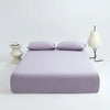 A purple ice silk cooling fitted bed sheet set is displayed on a white surface with a matching pillow and a white lamp.