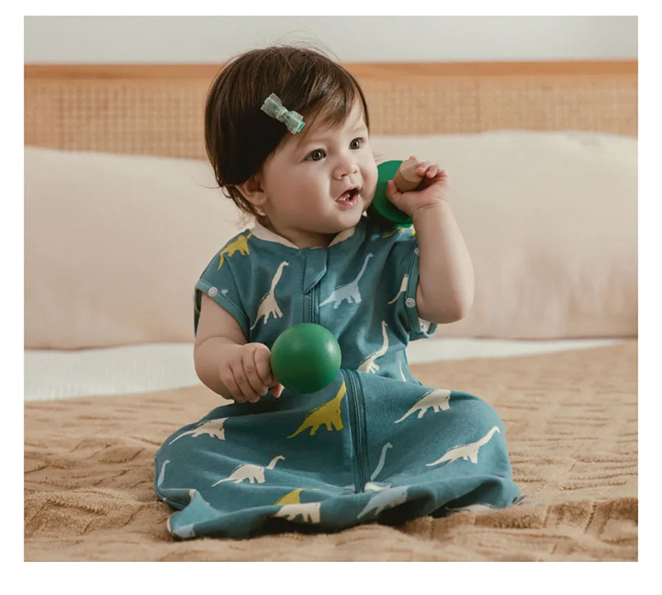 A baby wearing a teal dinosaur patterned sleeping bag is looking at a green ball.