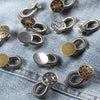 A collection of silver and brass flexible waist extender buttons for jeans and clothing laid out on a denim fabric.