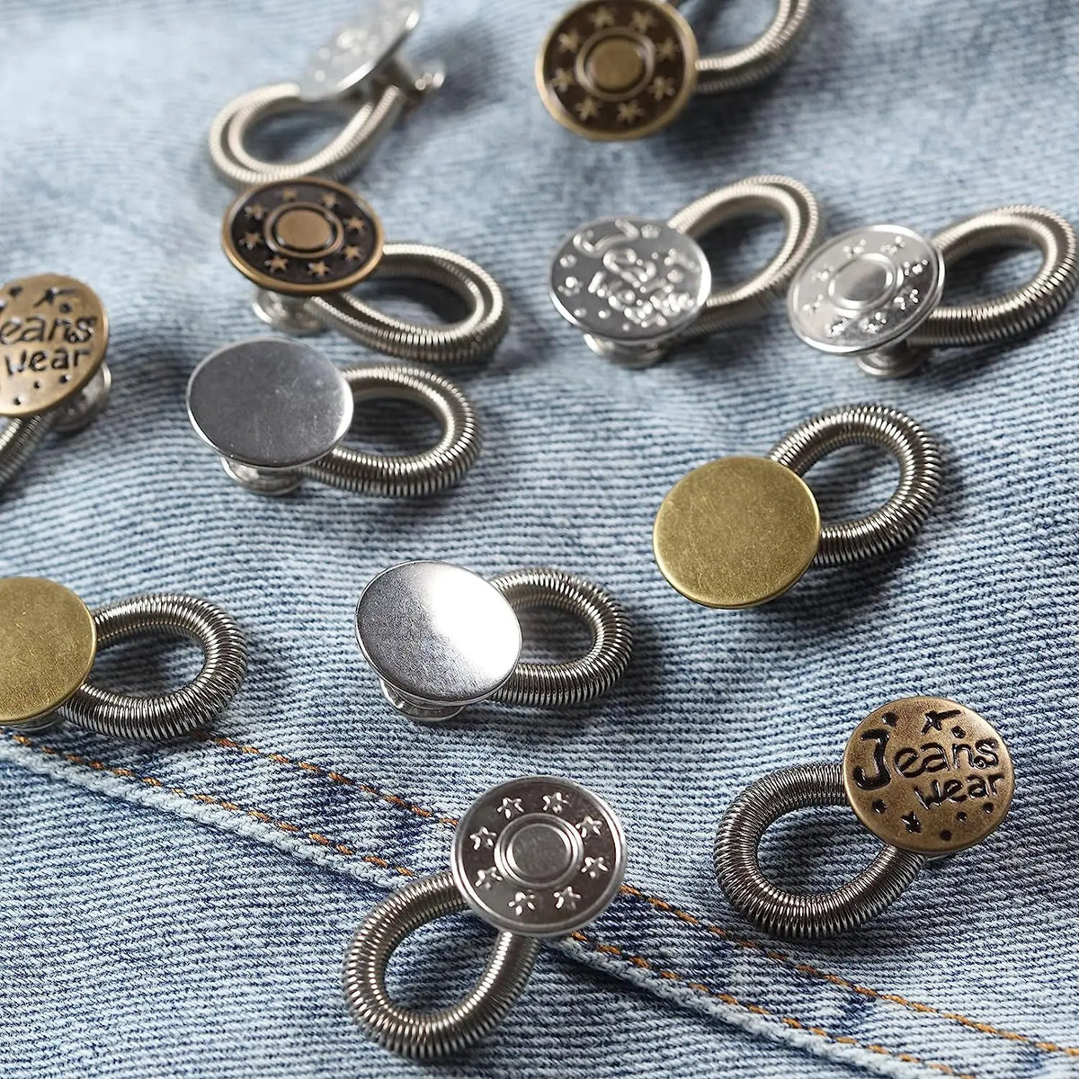 A collection of silver and brass flexible waist extender buttons for jeans and clothing laid out on a denim fabric.