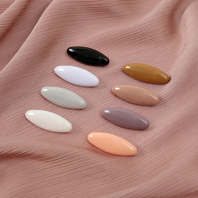 Eight plastic safety pins in various colors including black, white, beige, and pink, displayed on a pink fabric.
