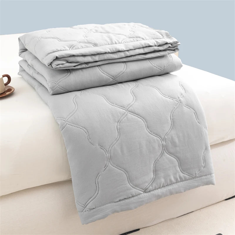 A stack of light gray quilted duvet covers and a folded duvet on a white bed.