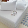 A white fitted bed sheet cover is displayed on a white bed with pillows and a small white clock.