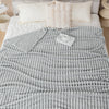 A gray knitted throw blanket with vertical ribbing is draped over a white bed, next to fluffy pillows.