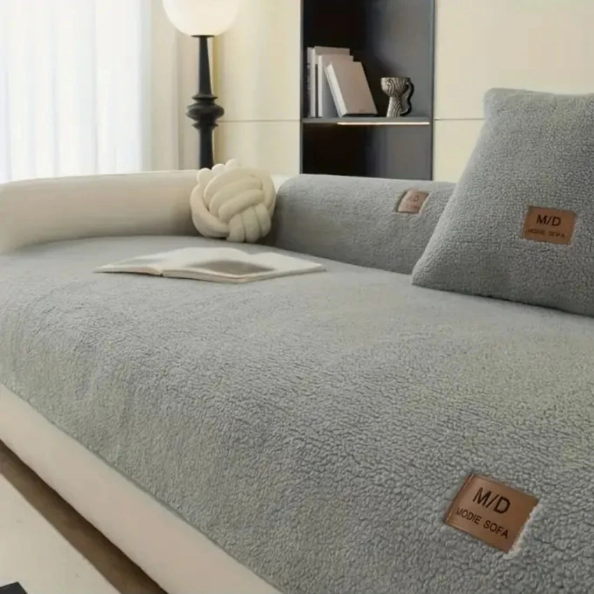 A light gray sherpa anti-slip sofa cover displayed on a sofa with a white cushion and decorative pillows.