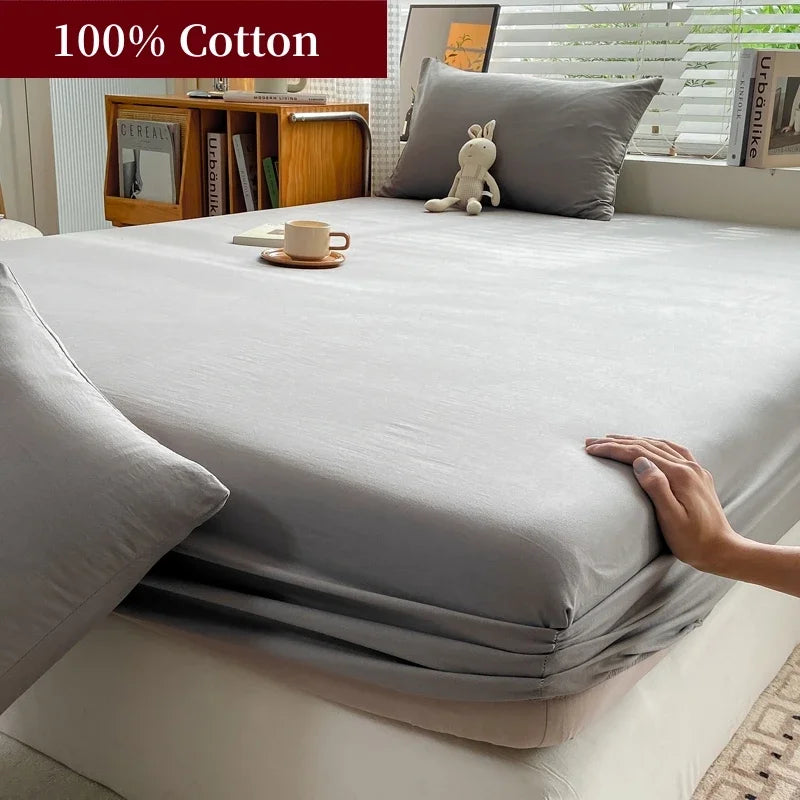 A fitted gray cotton bed sheet is draped over a bed, with pillows and wooden shelving visible in the background.