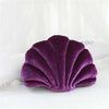 A deep purple velvet stuffed sofa cushion shaped like a decorative seashell resting on white fabric.