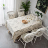 A dining table set with a botanical print linen tablecloth, white wooden chairs, and green foliage decor.