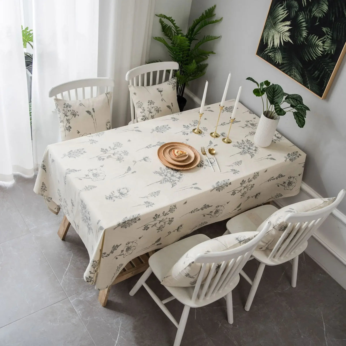 A dining table set with a botanical print linen tablecloth, white wooden chairs, and green foliage decor.