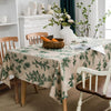 A botanical print linen tablecloth spread on a wooden table with white ceramics and greenery.