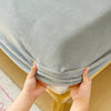 Hands are folding a light gray flannel fitted sheet with deep pockets on a wooden surface.
