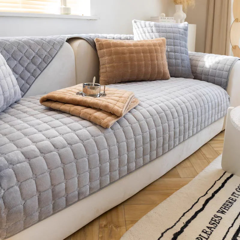 A light gray quilted sofa protection cover is displayed on a white sofa, with a tan throw blanket resting on it.