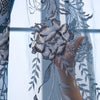 A hand is holding a detailed, silver-blue embroidered flower design on a sheer dual-layer curtain.