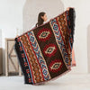 A woman holds up a dark brown and rust patterned Western checkered knit blanket with fringed edges.