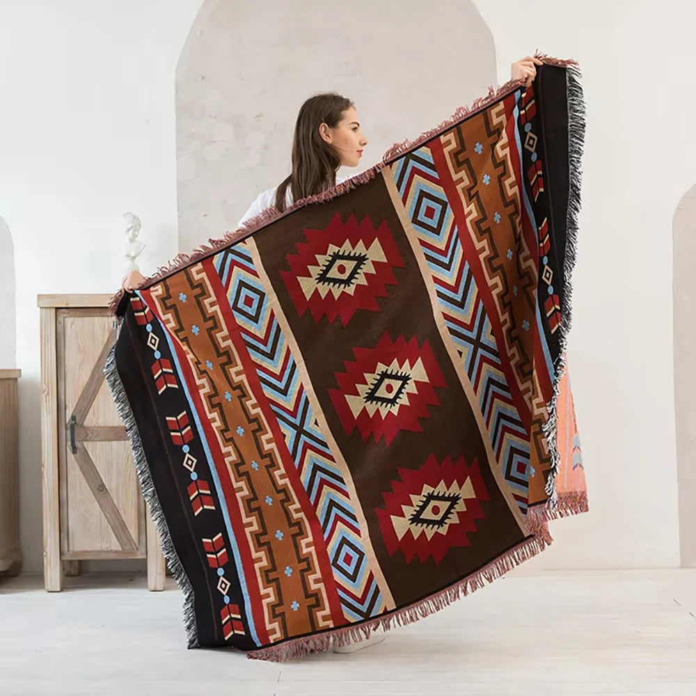 A woman holds up a dark brown and rust patterned Western checkered knit blanket with fringed edges.