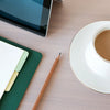 A wooden pencil rests on a pink surface next to a white cup and saucer, with a green notebook and a tablet visible.