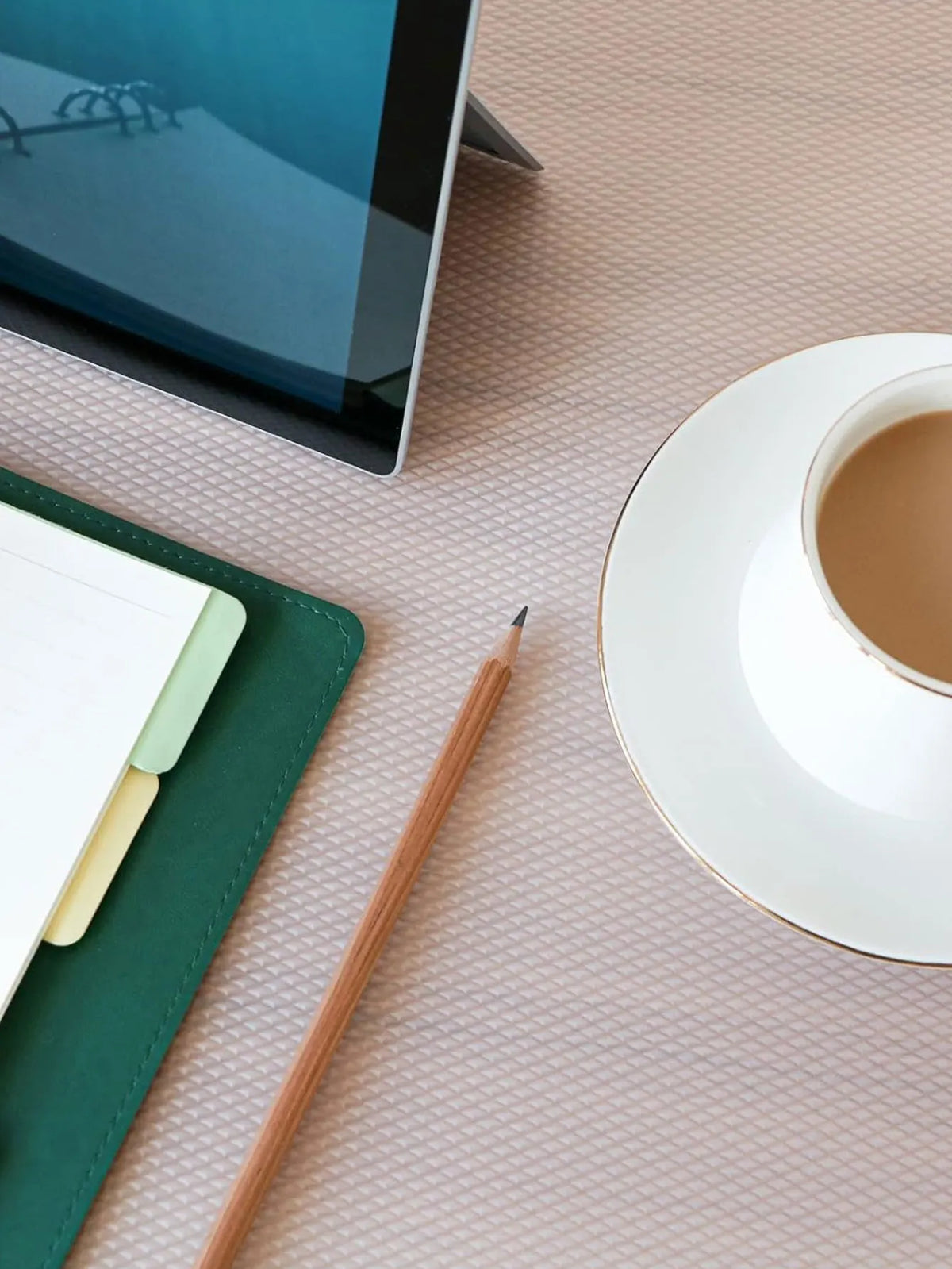 A wooden pencil rests on a pink surface next to a white cup and saucer, with a green notebook and a tablet visible.