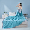 A woman models a teal grid quilt draped over a bed in a light blue room.