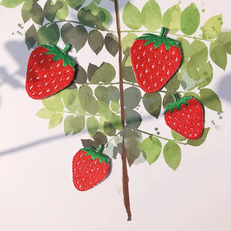 Three red strawberry embroidery patches are arranged on a green leaf background.