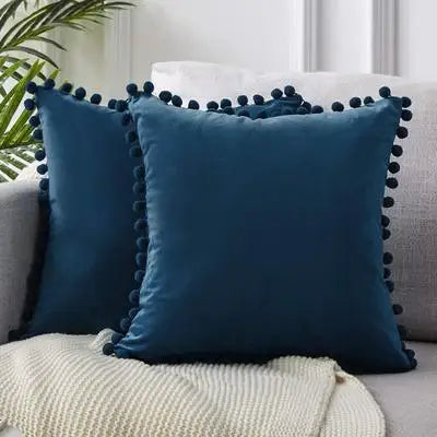A dark teal velvet decorative cushion cover with tasseled trim resting on a light fabric.