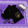 A black velvet tarot cards storage pouch with gold sun design and several tarot cards on a purple patterned surface.