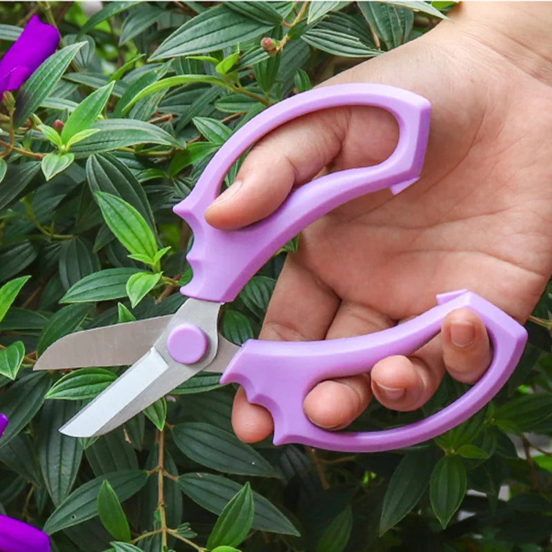A hand holds a purple precision bonsai and floral shears with a comfort grip against a green leafy background.