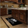 A black non-slip flannel area rug with white silhouette designs placed on a light wood floor in a kitchen setting.