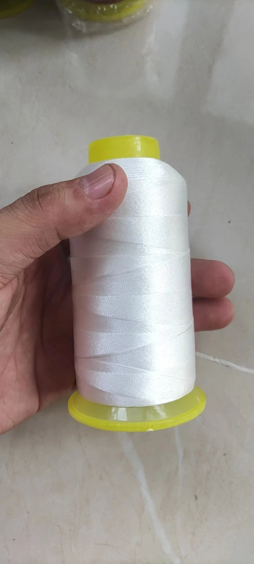 A hand is holding a spool of white high tenacity polyester sewing thread with yellow plastic caps.