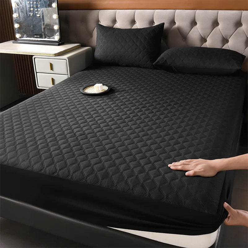 A black quilted mattress protector is shown on a bed with pillows and a bedside table.