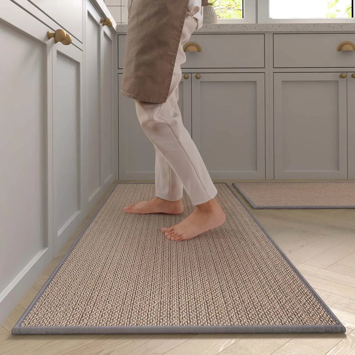 A person wearing light pants is standing on a rectangular beige non-slip faux sisal kitchen rug in a modern kitchen.