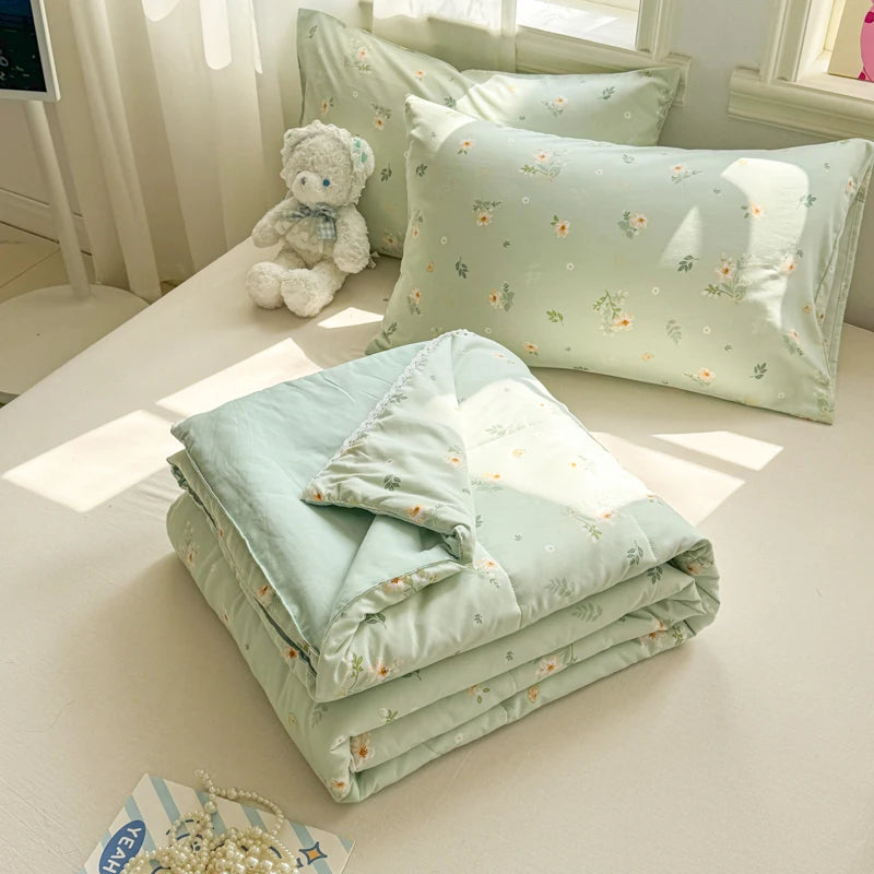 A light mint green quilted blanket is folded on a white surface next to several decorative pillows and a stuffed animal.