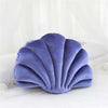 A purple velvet stuffed sofa cushion shaped like a decorative seashell resting on white bedding.