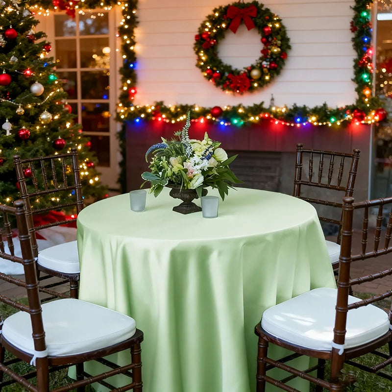 A round table set for an event with a light green satin overlay cloth, surrounded by wooden chairs in front of a decorated wa