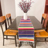 A dark wooden dining table set with a colorful cotton Mexican serape table runner and a vase of red flowers.