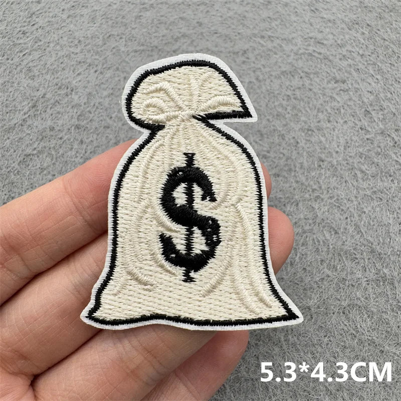 A beige embroidered iron-on patch shaped like a money bag with a dollar sign on it being held in a hand.