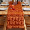 An orange textured table runner decorated with carved pumpkins and small gourds on a wooden dining table.